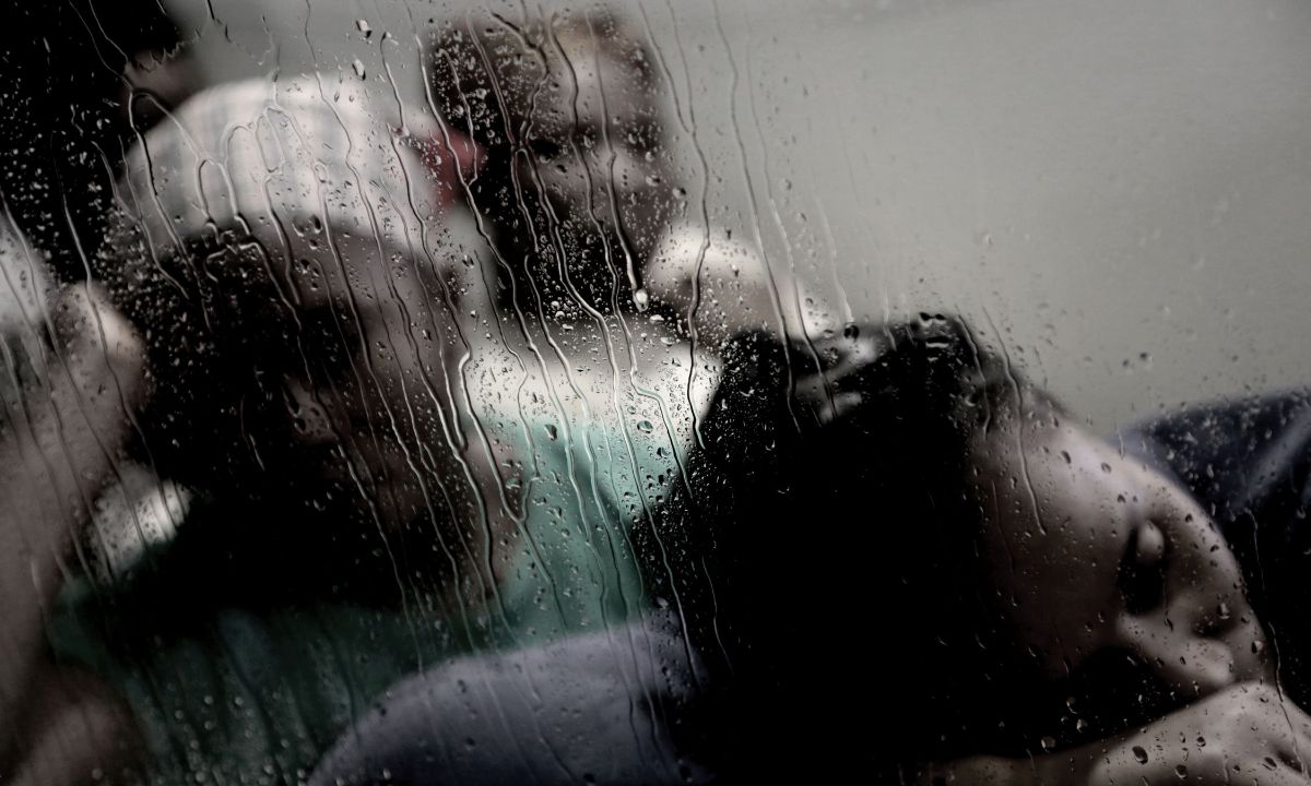 Drei Frauen sitzen hintereinander. Die Vorderste legt ihren Kopf auf ihren Arm ab, die zweite stützt ihren Kopf auf der rechten Hand auf. Dahinter ist das Gesicht einer dritten Frau zu erkennen. Ihre Gesichter sind unscharf, sie befinden sich hinter einer nassen Glasscheibe.