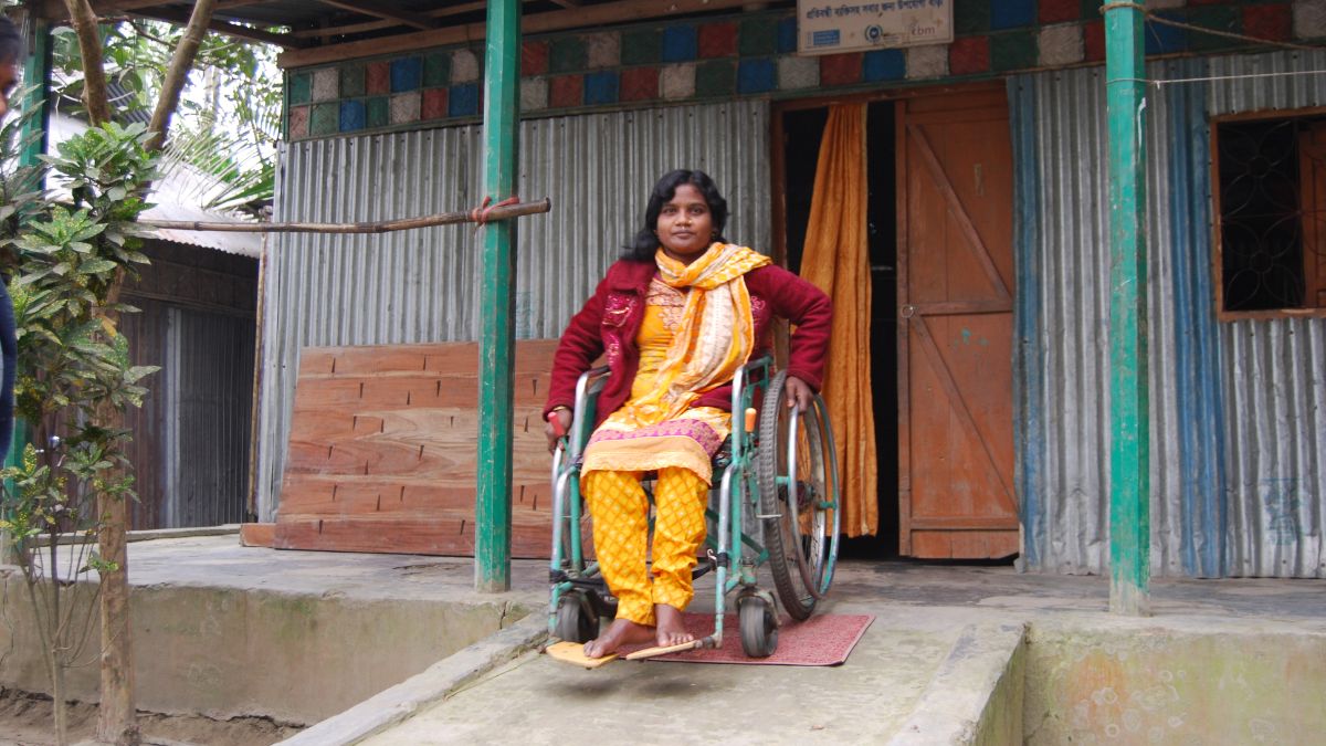 Eine junge Frau aus Bangladesch sitzt im Rollstuhl. Dieser steht auf einer vor einem Haus angebrachten Rampe steht.