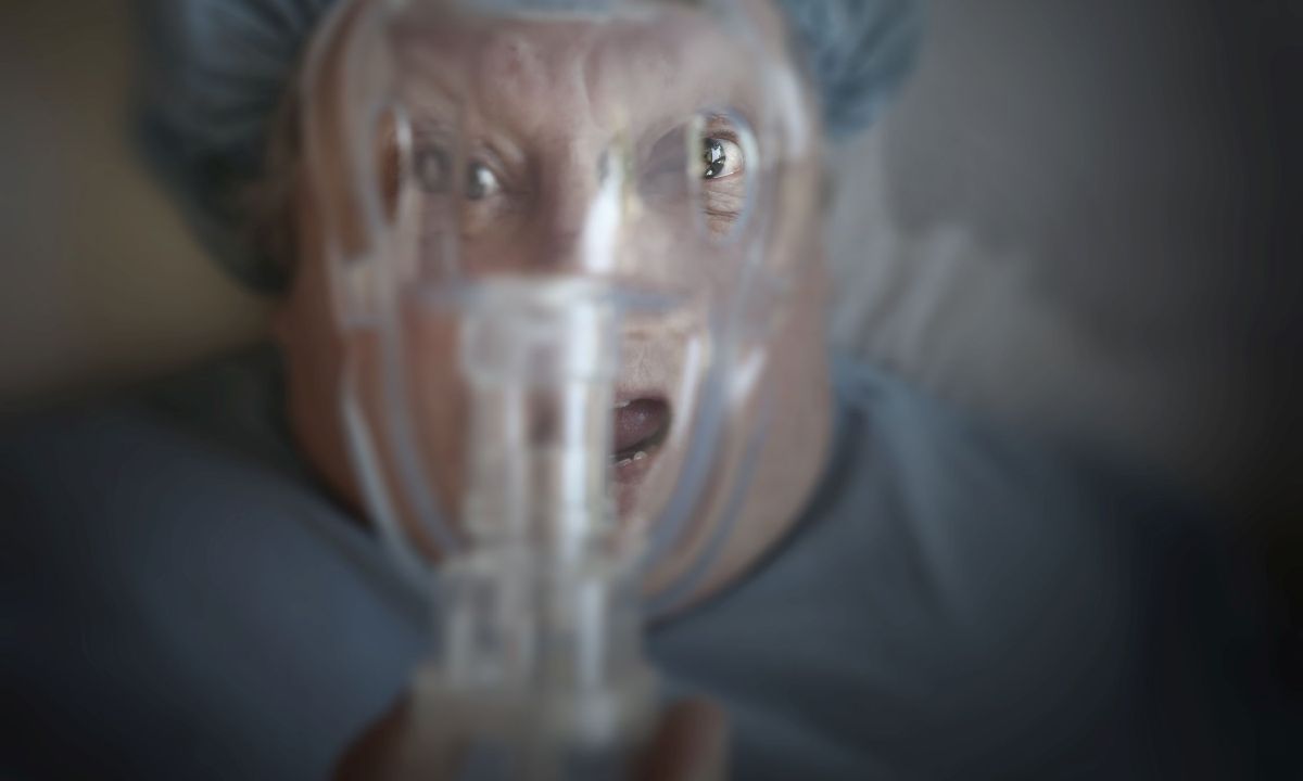 Eine Frau mit aufgrissenen Augen und offenem Mund in OP-Kleidung schaut durch eine Narkosemaske vor ihrem Gesicht.