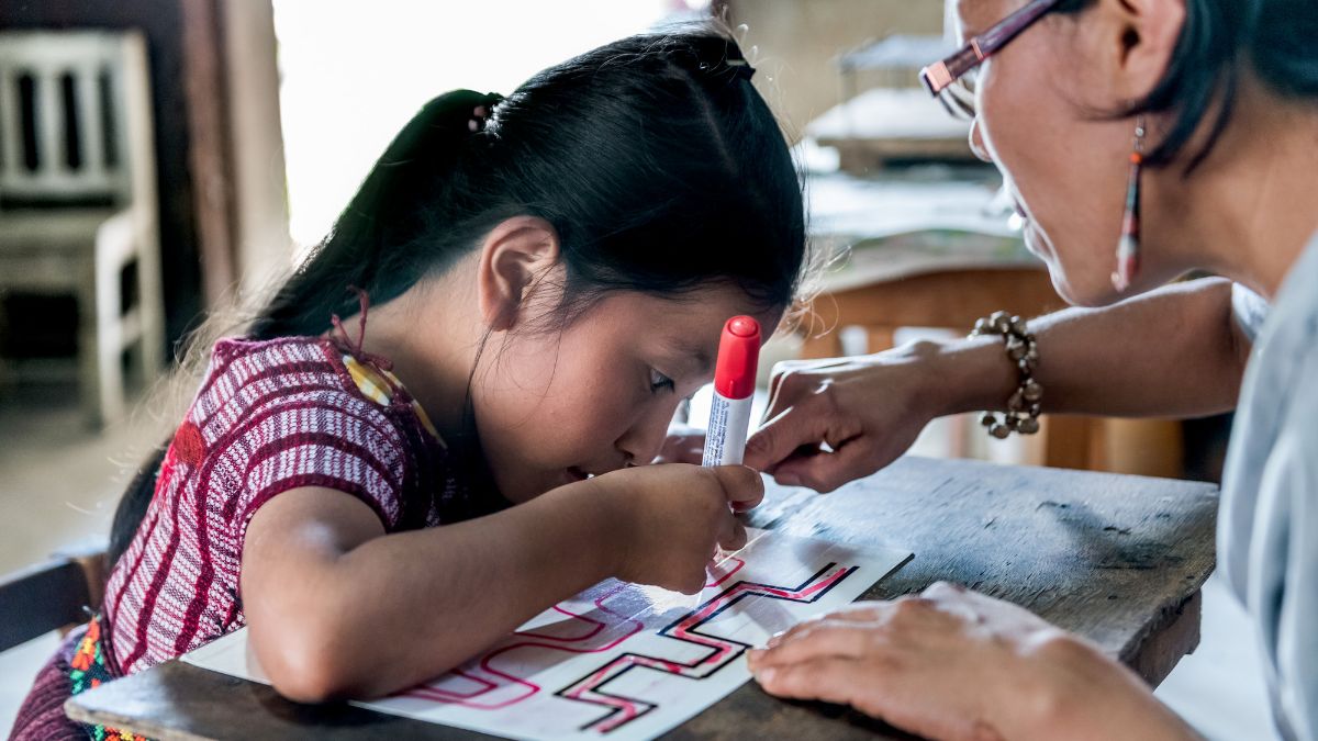 A child drawing on a piece of paper. A woman is sitting with her.