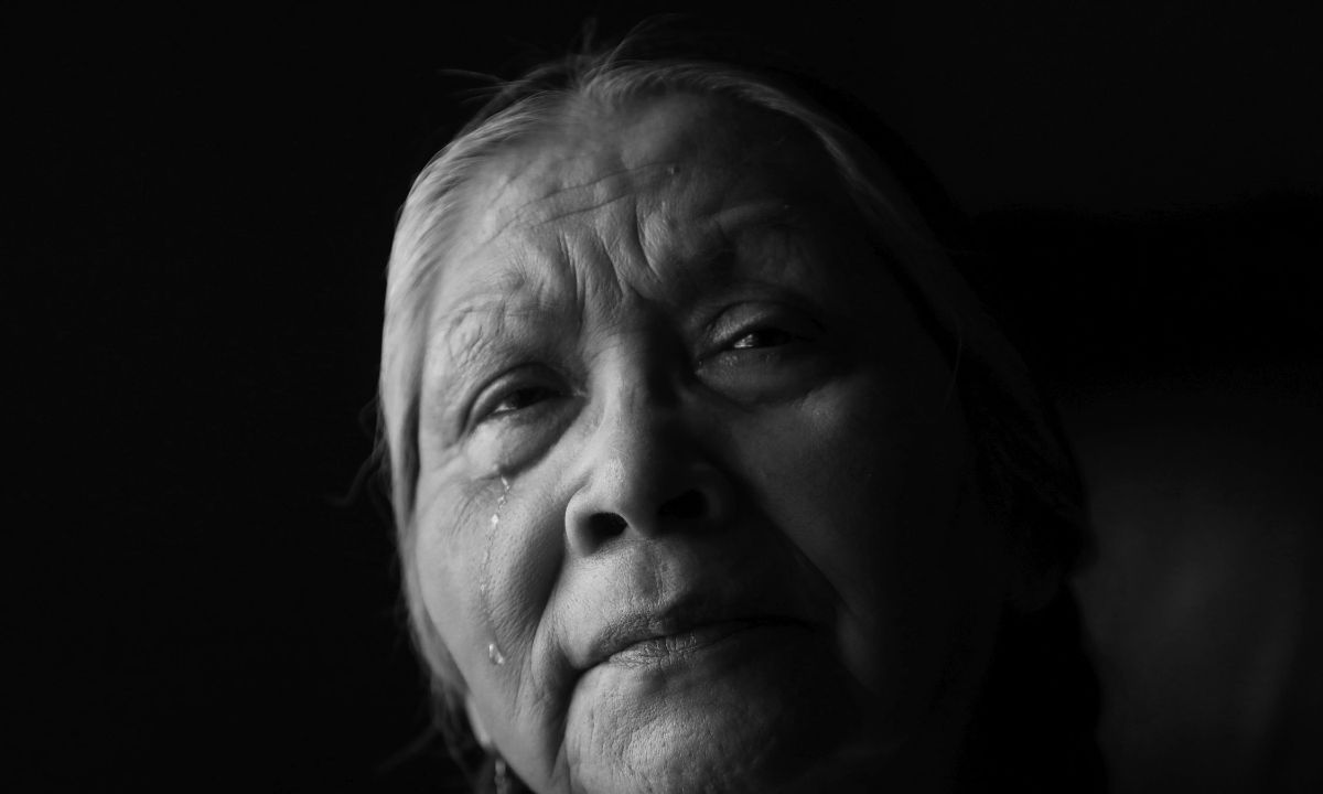 Gesicht einer älteren, indigenen Frau. Sie blickt ernst und runzelt die Stirn, eine Träne läuft ihr die Wange herunter.