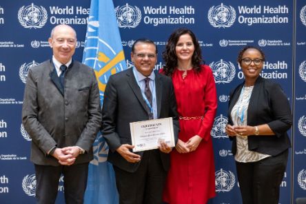 Four people standing in front of a backdrop featuring the World Health Organization (WHO) logo. One of the individuals at the centre holds a certificate that reads, &quot;Certificate of Membership&quot;, awarded to the Christian Blind Mission (CBM). The people in the photo are dressed in formal attire. 